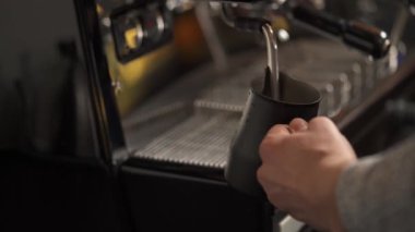 Sütlü sürahiyi tutan bir elin yan görüntüsü ve kahve makinesinde ısıtan bir buhar çubuğu. Barda kahve yapmak.