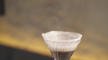 A close-up of a kettle and a spout with hot water poured into filter with coffee. Water is dripping through filter of the pour over kit and it is steaming a bit. Coffee making in a bar