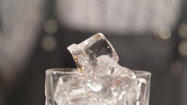 A close-up of a drinking glass with ice cubes and a pipette above it dropping brown liquid cocktail bitter drops.