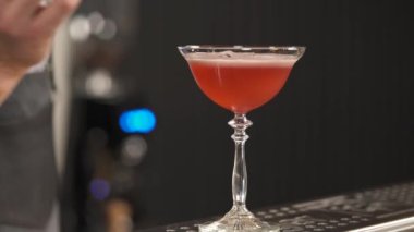 A tumbler with pink liquid on the bar counter and a hand putting a rosemary sprig on the top