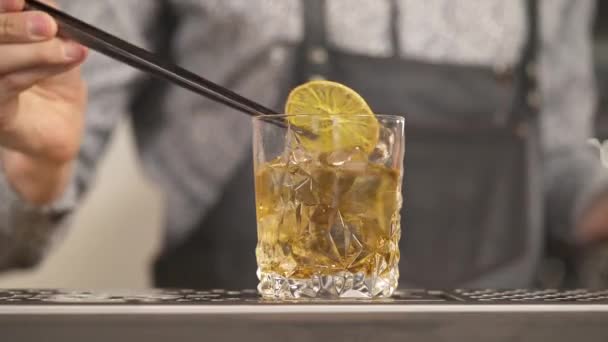 氷と茶色の液体と手がピンセットを保持し 上にレモンスライスを置くとガラスのクローズアップ バーでのドリンクの準備 — ストック動画