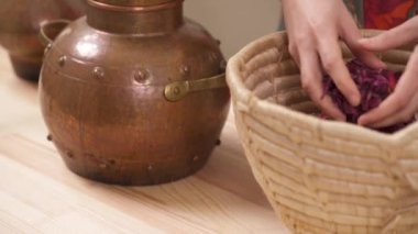 A close-up view of woman's hands filling copper alembic with dried rose petals taken from a wicker basket.