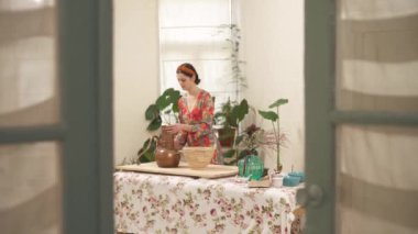 A view of woman filling alchemical still with dried rose petals taken from a wicker basket. Approaching camera