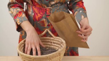 A close-up view of woman's hand holding the paper package. With her other hand woman is filling it with dried lavender flowers by taking them from a wicker basket.