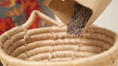 A close-up view of a wicker basket filling by dried lavender flowers from the paper package on the blurred background.