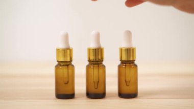 A close-up front view of woman's hand choosing central small amber bottle with white cap out of three bottles standing on the wooden table.