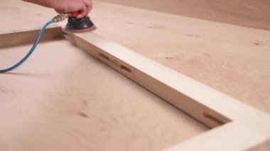 A front view of man's hand holding a pneumatic power sander and polishing a wooden frame joined by bridle joint