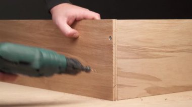 A front view of man's hand holding cordless electric screwdriver and making a hole in a plywood frame