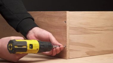 A front view of man's hand holding an electric screwdriver and screwing a bolt into plywood boards