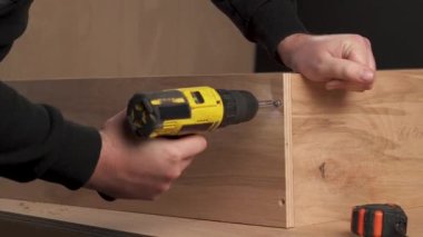 A front view of man's hand holding electric screwdriver and screwing two bolts into holes in plywood box