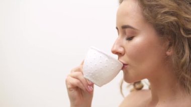 A view from side of a cheerful young woman with curly hair drinking from white cup and smiling on white background
