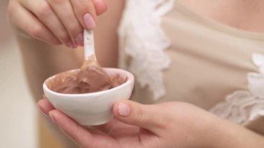 A close-up of a young woman mixing the ingredients of the natural brown mask in a ceramic dish and testing it on the skin of her hand