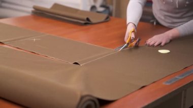 A tailor cutting a piece of large skein of brown fabric with professional scissors according to the pattern