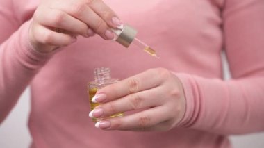 A closeup of woman's hand opening bottle with natural oil, filling a pipette and applying oil on the back side of her palm. She is gently rubbing oil into the skin