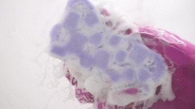 Demonstration close-up video of woman's hand in pink rubber glove squeezing soapy colorful sponge while cleaning glass surface. Clean house conception