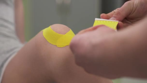 理学療法士は 女性患者の膝にKinesioテープを置きます 膝関節治療 スポーツ及びリハビリテーション — ストック動画
