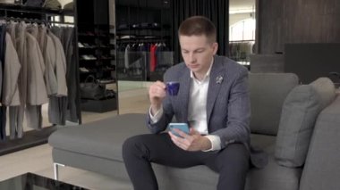 Yakışıklı müşteri, lüks bir erkek giyim mağazasında kahve içerken akıllı telefonunun ekranına bakıyor..