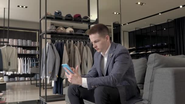 年轻人坐在灰色时髦的沙发上 看着他的智能手机屏幕上的男性服装店 男性时尚精品店 商店和销售概念 — 图库视频影像