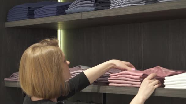 男性の洋服屋さんの棚に 明るくスタイリッシュな男性用Tシャツを丁寧に置いたお店のアシスタントのクローズアップ ファッション店のルーティンワーク — ストック動画