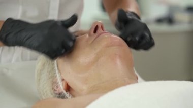 Lüks kaplıca salonunda yüz masajı yaptırırken dinlenen çekici bir son sınıf kadını, yakın plan. Kozmetik uzmanı yüz masajı yapıyor.