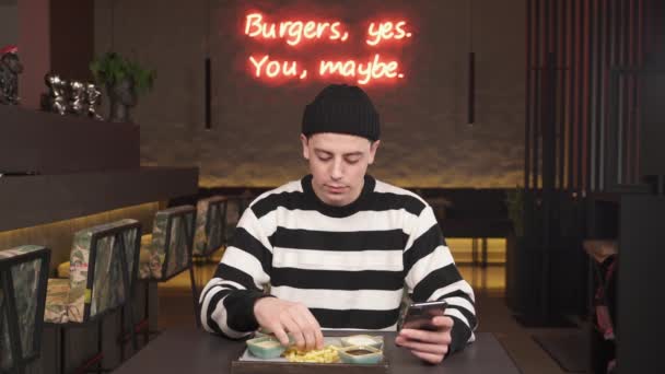 フランスのフライドポテトを食べながら 現代的なレストランに座っている間 集中した若い男が保持し 電話を見て — ストック動画