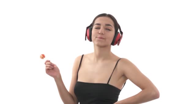 無線のイヤホンで音楽を聴き 甘いロリポップを食べる若い女性のクローズアップショット 屈託のない女踊りの肖像 — ストック動画