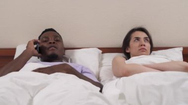 Yatakta melez bir çiftin portresi. Beyaz kız Afrikalı erkek arkadaşına kızıyor çünkü adam akıllı telefondan konuşmayı kesmiyor.