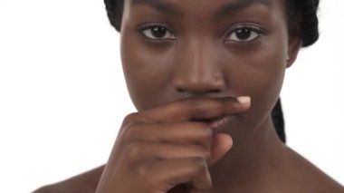Genç Afrikalı bir kadının yakın plan portresi. Parmağını ağzının üstünde yatay olarak tutuyor ve sağa sola vuruyor. Burnum kaşınıyor. Beyaz arkaplanda izole