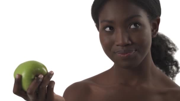 緑のリンゴを食べるかどうか疑問に思う若いアフリカ人女性の肖像画を閉じ 最終的に白を背景に喜びでそれを食べる — ストック動画