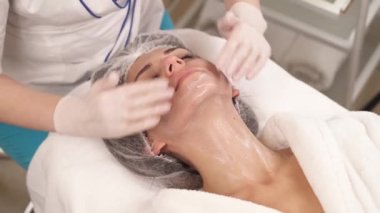 Profesyonel kozmetik uzmanı yüz kremi sürerken ve kaplıca salonunda yüz masajı yaparken gözleri kapalı koltukta yatan bir kadının yakın plan görüntüsü. Cilt bakımı kavramı