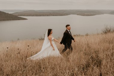 Beyaz elbiseli ve takım elbiseli, sevgi dolu bir çift nehrin yukarısındaki dağdaki sonbahar tarlasında uzun çimlerin üzerinde öpüşüyor. Gün batımı. bakota ukraine