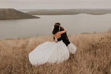 Beyaz elbiseli ve takım elbiseli, sevgi dolu bir çift nehrin yukarısındaki dağdaki sonbahar tarlasında uzun çimlerin üzerinde öpüşüyor. Gün batımı. bakota ukraine