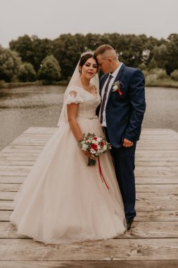 Sarışın, Avrupalı, beyaz erkek, mavi takım elbiseli damat ve siyah saçlı, uzun peçeli, başında taç olan gelin. Yeni evliler, gölde tahta iskele köprüsünde öpüşüyorlar.