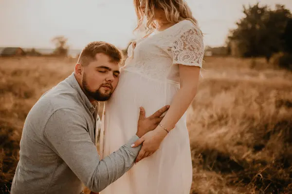 husband hugs pregnant wife father listens belly put his ear to tummy. Happy family resting in nature hugs kisses in summer at sunset. future mother Caucasian woman in white cotton dress