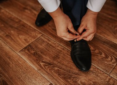 Beyaz gömlekli ve siyah pantolonlu bir adam, kahverengi desenli, ahşap bir zeminde siyah botlarının bağcıklarını bağlayan bir sandalyede oturuyor. Erkekler klasik moda resmi ayakkabıları..