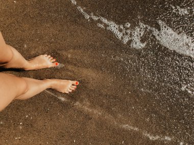 Yaz günü kumsalda, deniz köpüğü dalgalarında çıplak ayaklı bir kadın. Kadın ayağının üstündeki üst manzara. Okyanus berrak deniz köpüğü. Sahilde yaz tatili konsepti olan bir kız. Doğal duvar kağıdı