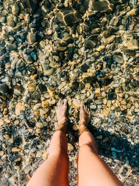 Woman legs barefoot at sea waves stones shells beach summer day. top view above women feet. Ocean clear water sea. girl on beach Summer background nature. top sea wallpaper Two feet dipping in water