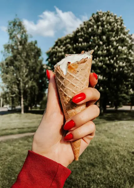 Yazın güneşli bir günde elinde dondurma tutan kadın. Kapatın. Doğal yeşil arka plan. Kırmızı manikürlü kız tırnakları. Waffle bardağındaki dondurma külahları. Yaz arkaplanı. Dondurmayı elle tutan kadın.