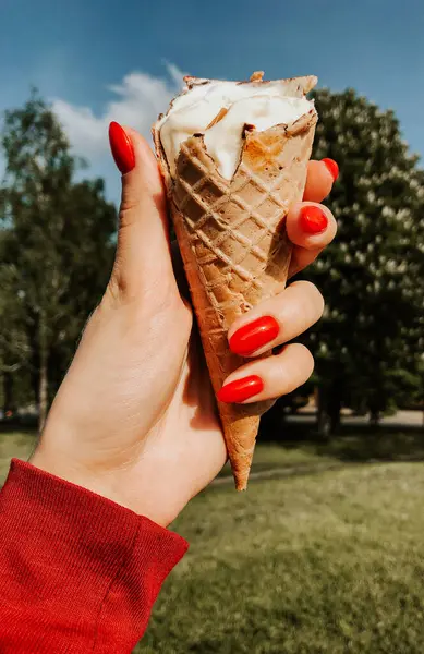 woman hand holding ice-cream at summer sunny day close up. natural green background. red manicure girl nails. ice cream cones in a waffle cup. summer background. woman holding the ice cream by hand