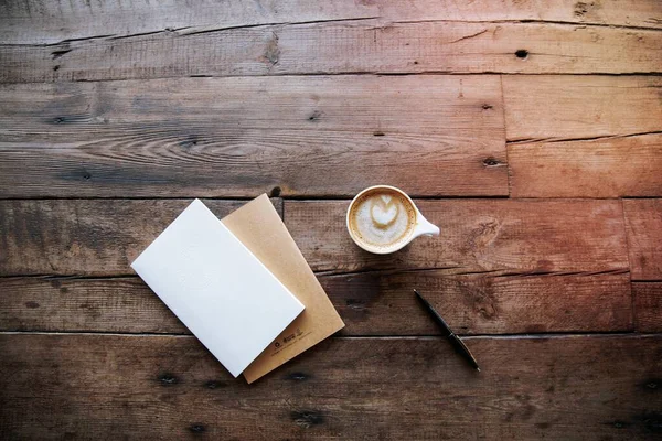 An overhead shot of a cup of coffee, a pen and two notebooks on a wooden surface.