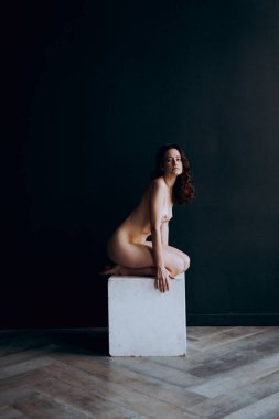Beautiful, natural naked girl artistic posing at a stylish and fashionable nude photo shoot as a model in a photo studio with a transparent cape and bandage and looks like art