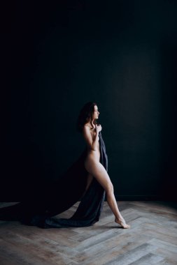 Beautiful, natural naked girl artistic posing at a stylish and fashionable nude photo shoot as a model in a photo studio with a transparent cape and bandage and looks like art