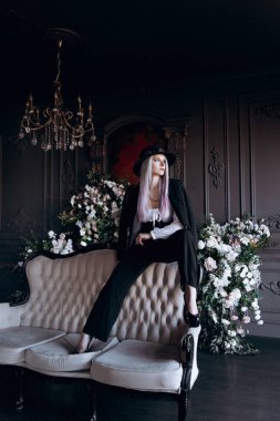 Luxurious girl with beautiful hair and tattoos in a hat and with a mouthpiece in a photo studio in a trouser suit.