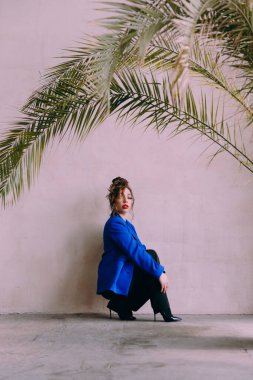 Güneş gözlüklü, ince vücutlu, mavi ceketli, yeşil elbiseli, şık, şık bir moda çekiminde fotoğraf stüdyosunda poz veren çekici bir kız..