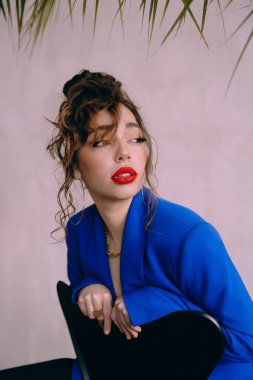 Güneş gözlüklü, ince vücutlu, mavi ceketli, yeşil elbiseli, şık, şık bir moda çekiminde fotoğraf stüdyosunda poz veren çekici bir kız..