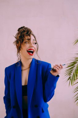 Güneş gözlüklü, ince vücutlu, mavi ceketli, yeşil elbiseli, şık, şık bir moda çekiminde fotoğraf stüdyosunda poz veren çekici bir kız..