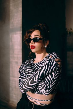 Güneş gözlüklü, ince vücutlu, mavi ceketli, yeşil elbiseli, şık, şık bir moda çekiminde fotoğraf stüdyosunda poz veren çekici bir kız..