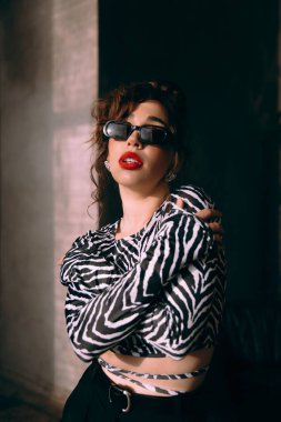 Güneş gözlüklü, ince vücutlu, mavi ceketli, yeşil elbiseli, şık, şık bir moda çekiminde fotoğraf stüdyosunda poz veren çekici bir kız..