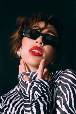 Güneş gözlüklü, ince vücutlu, mavi ceketli, yeşil elbiseli, şık, şık bir moda çekiminde fotoğraf stüdyosunda poz veren çekici bir kız..