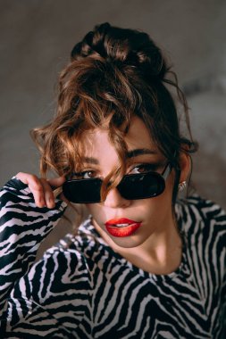 Güneş gözlüklü, ince vücutlu, mavi ceketli, yeşil elbiseli, şık, şık bir moda çekiminde fotoğraf stüdyosunda poz veren çekici bir kız..